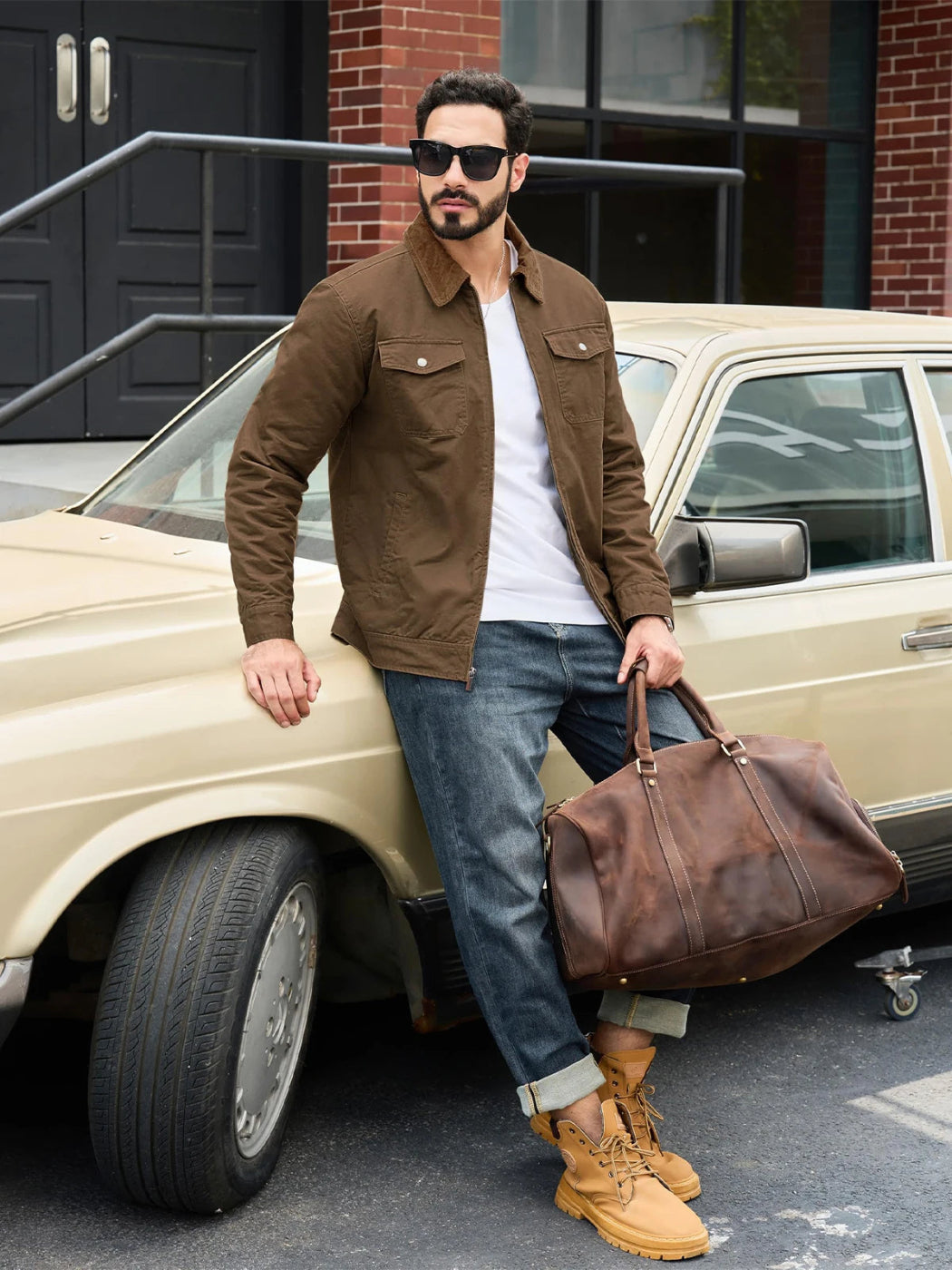 Man in brown jacket and jeans holding a leather bag next to a vintage car.